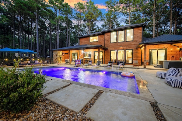 A two-story wooden house with a swimming pool, outdoor seating, and a view of the surrounding forest.