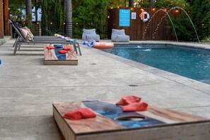 A poolside area with lounge chairs, a wooden table with red cushions, and a sign indicating pool rules. - Resort Pool & Hot Tub | Arcade + Bunk Room (Broken Bow)