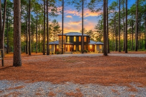 A modern two-story house surrounded by tall pine trees with a gravel driveway. - Resort Pool & Hot Tub | Arcade + Bunk Room (Broken Bow)