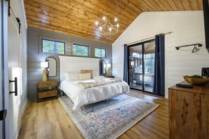 A bedroom with a large bed, a chandelier, a nightstand, a mirror, and a view through a glass door.
