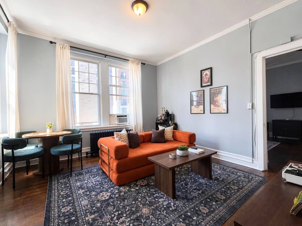 Ein Wohnzimmer mit einer Couch, einem Couchtisch und Sesseln. Es gibt einen Esstisch mit Stühlen, ein Fenster mit Vorhängen und einen Fernseher.