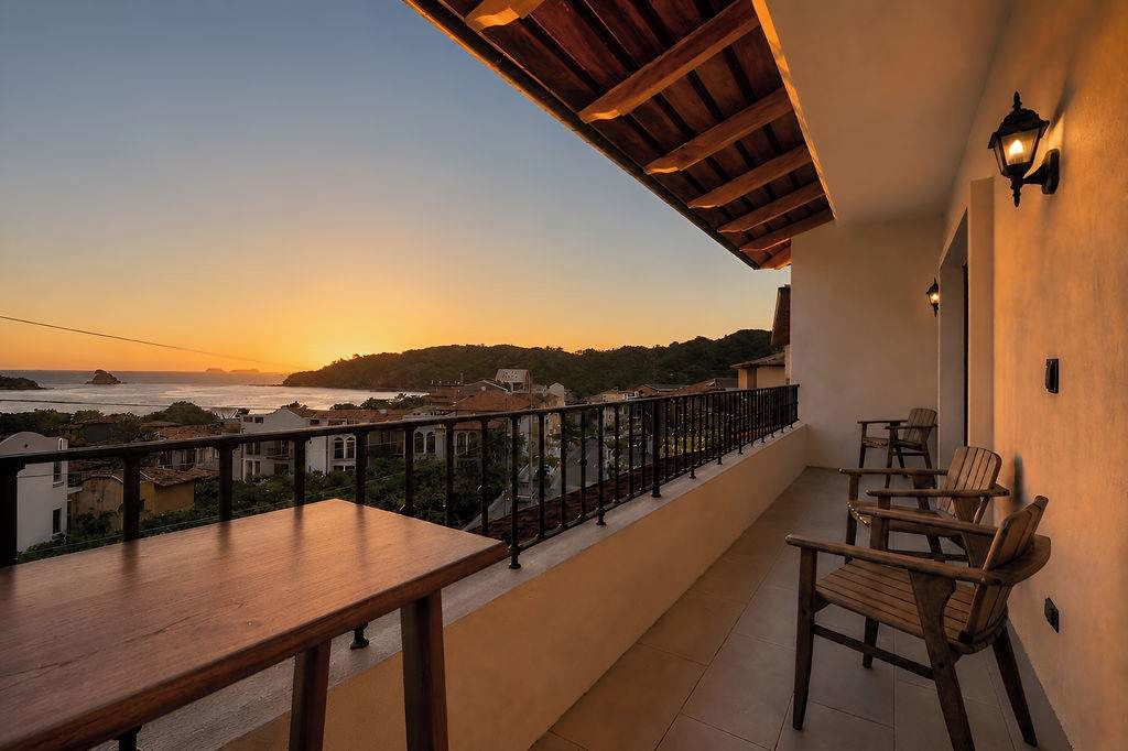 Un balcón con vista al mar y edificios al atardecer. Se observan sillas y una mesa de madera.