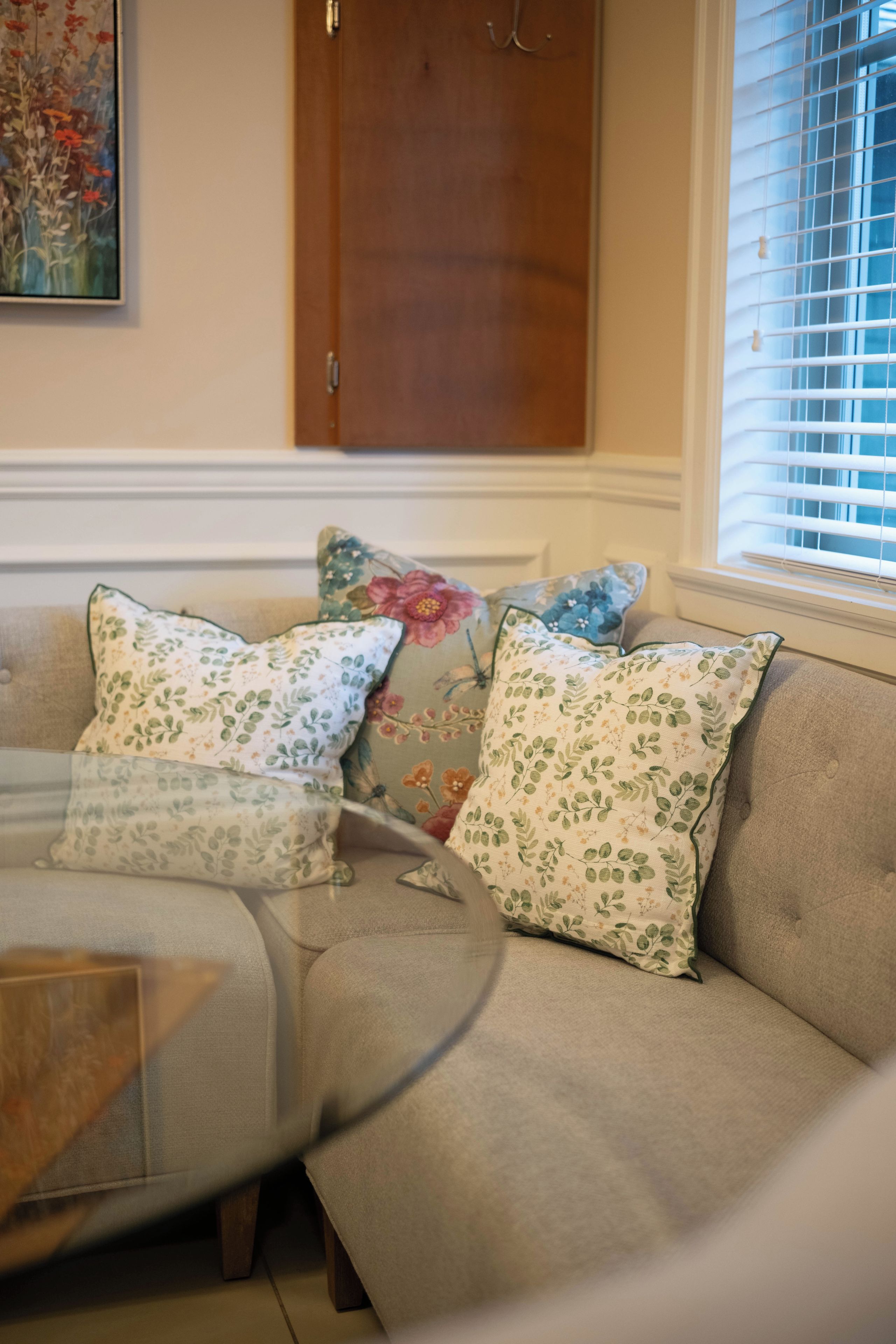 A beige sofa with floral-patterned cushions, a wooden door, and a window with blinds.