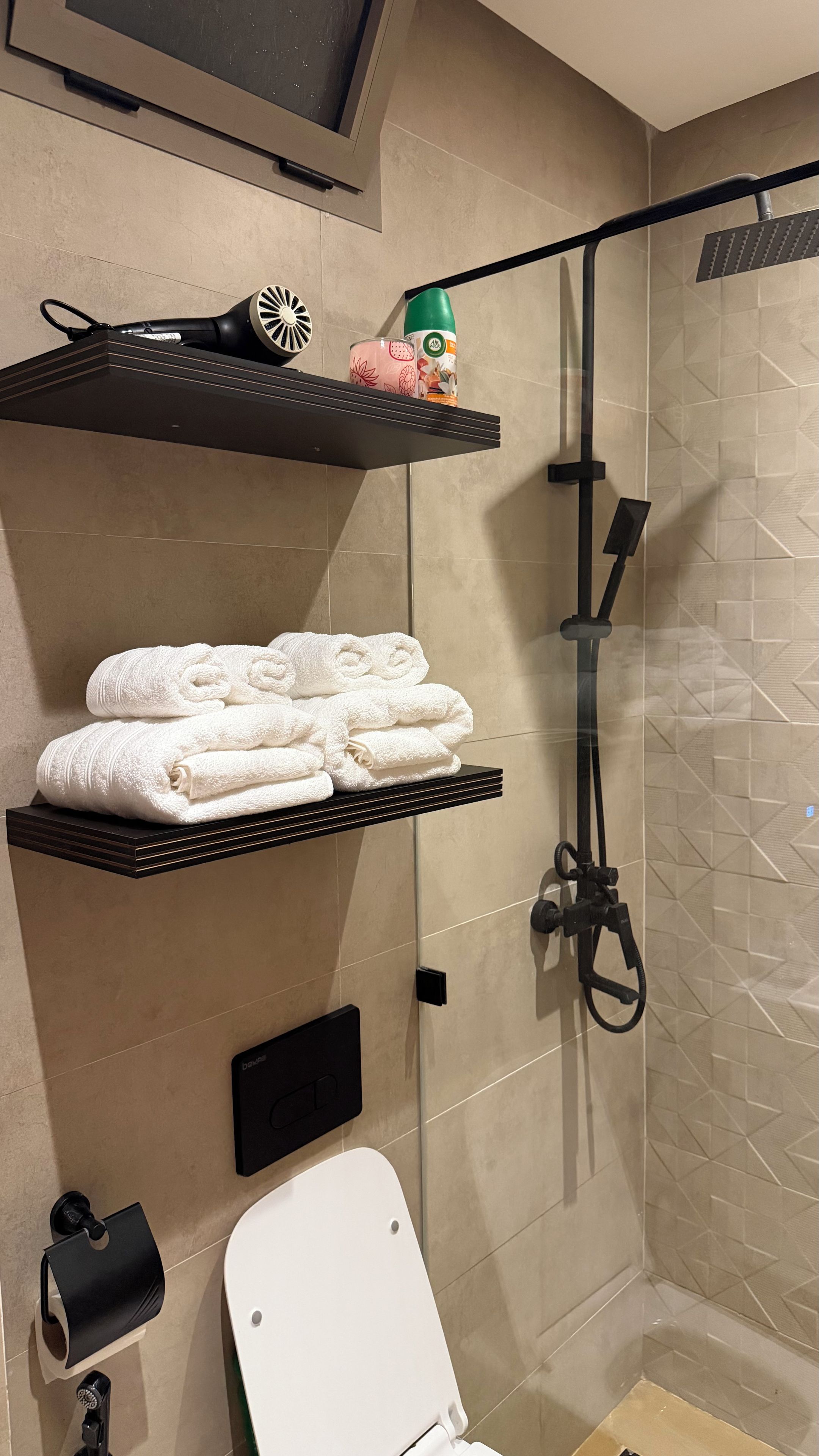 A bathroom with a shower, a toilet, and shelves with towels and a hairdryer.