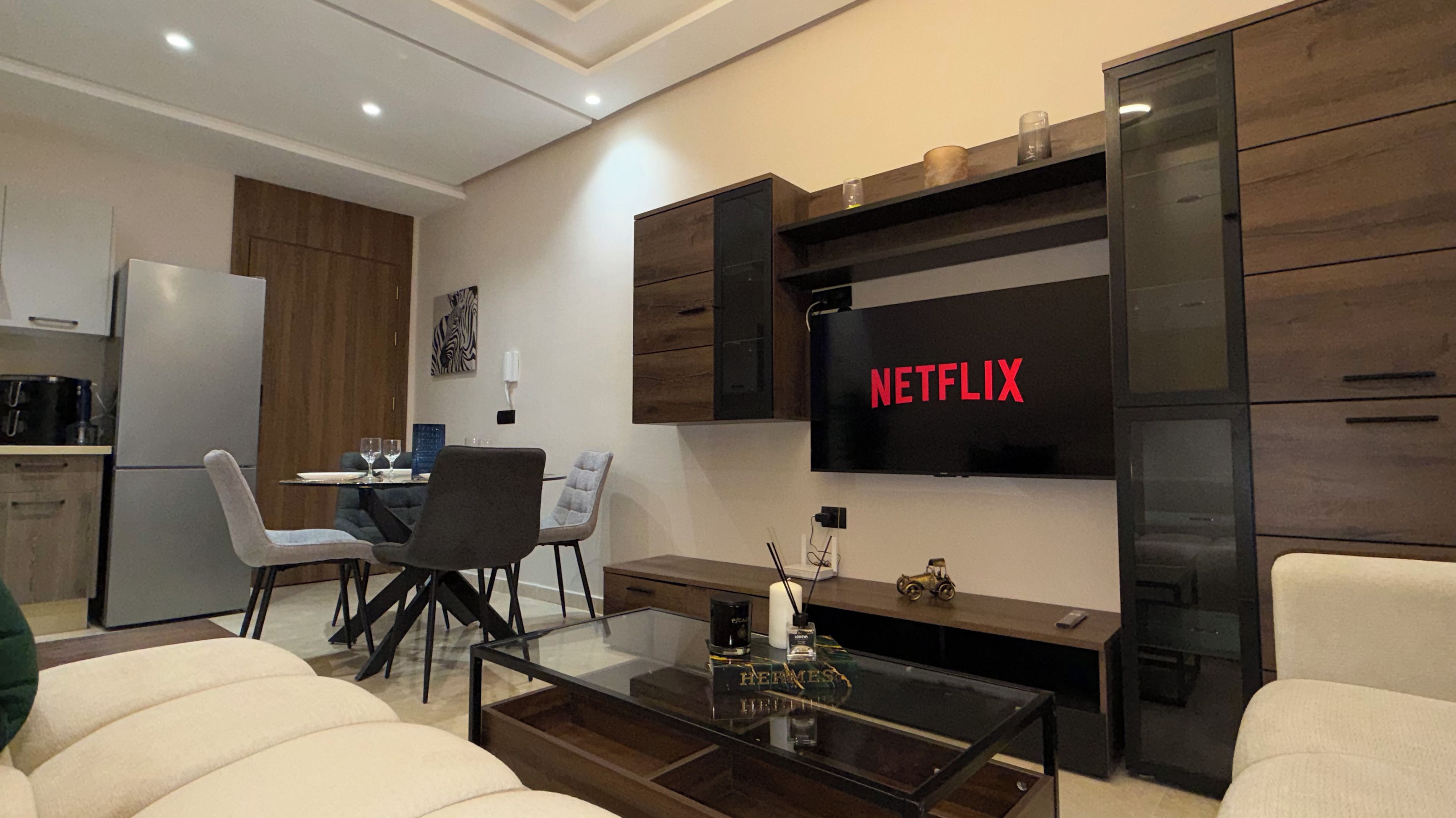 Un salon moderne doté d’un téléviseur à écran plat fixé au mur, d’un coin repas avec des chaises et une table, et d’une cuisine équipée d’un réfrigérateur.