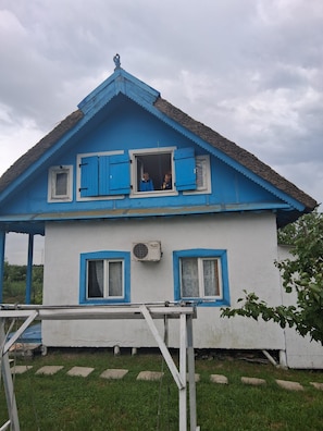 A two-story house with a blue roof and white walls, featuring two windows with shutters and a visible air conditioning unit. - Terrific 3-bedroom house in enchanting Mila 23 awaits you (Mila 23)