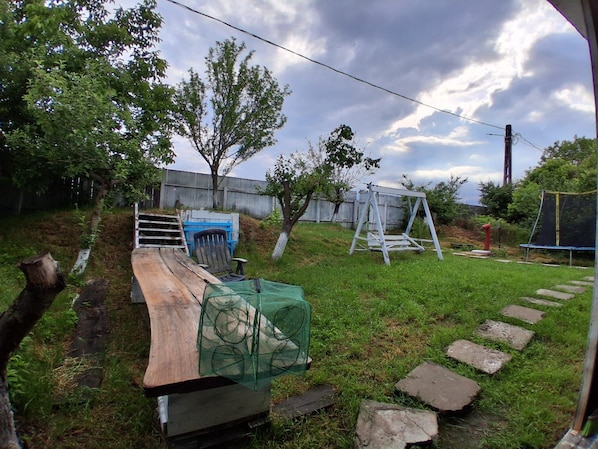 A backyard with a wooden table, a blue chair, a green netted object, a swing set, and a trampoline.