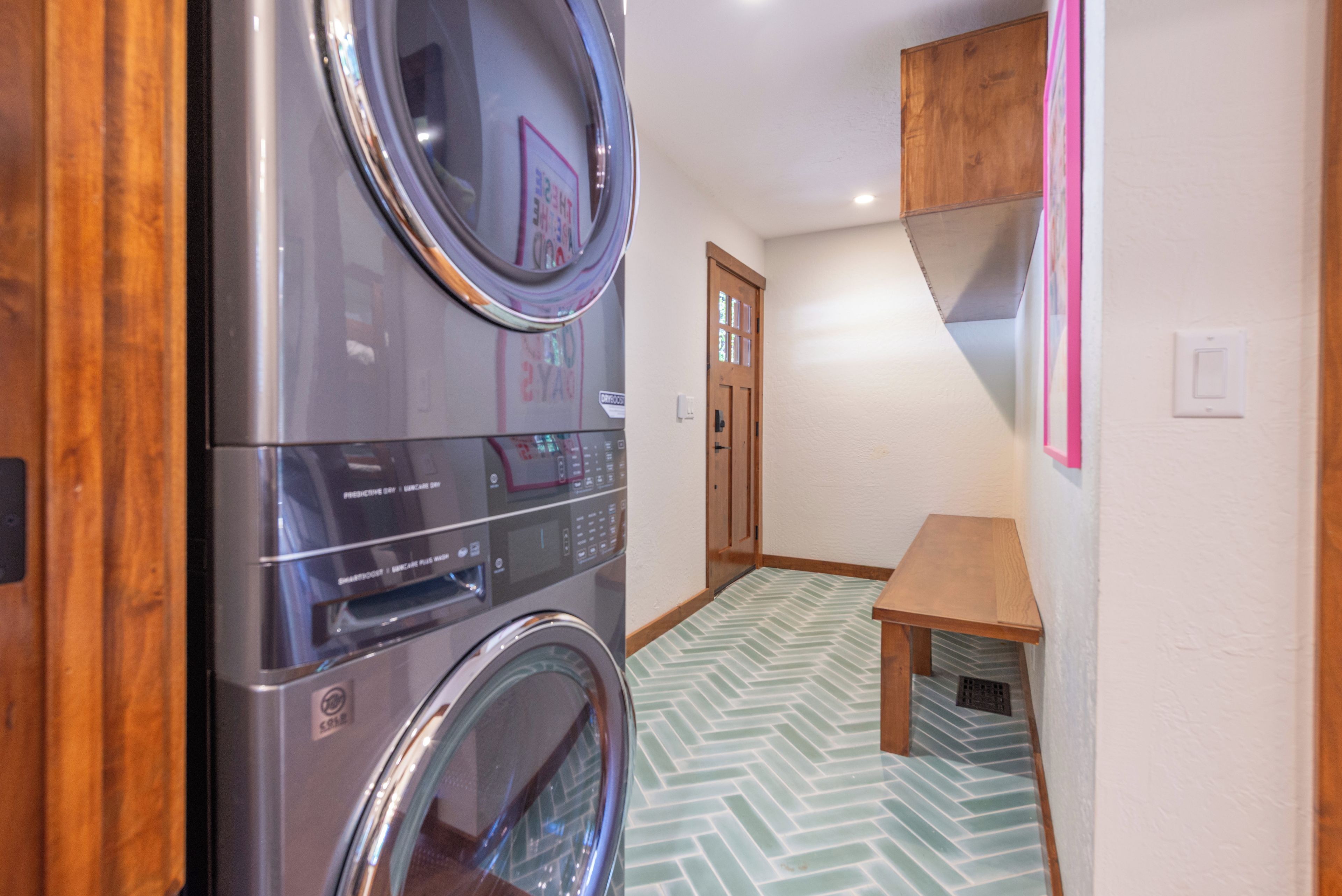 A laundry room with a front-loading washing machine and dryer, a wooden bench, and a door leading to another room.