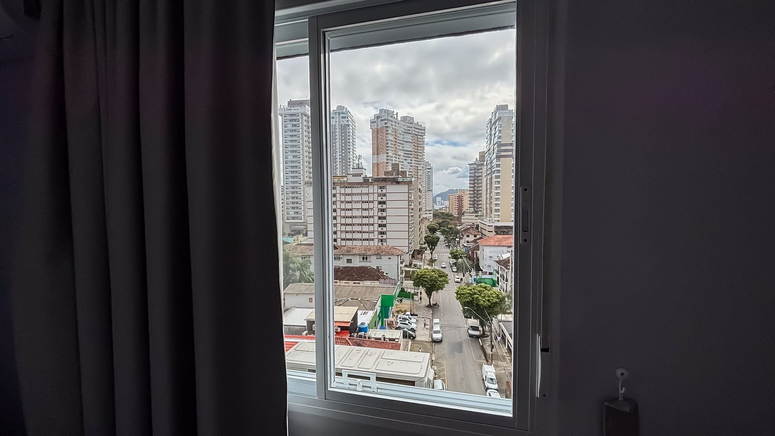 A view through a window of a cityscape with tall buildings, a street with parked cars, and a green structure.