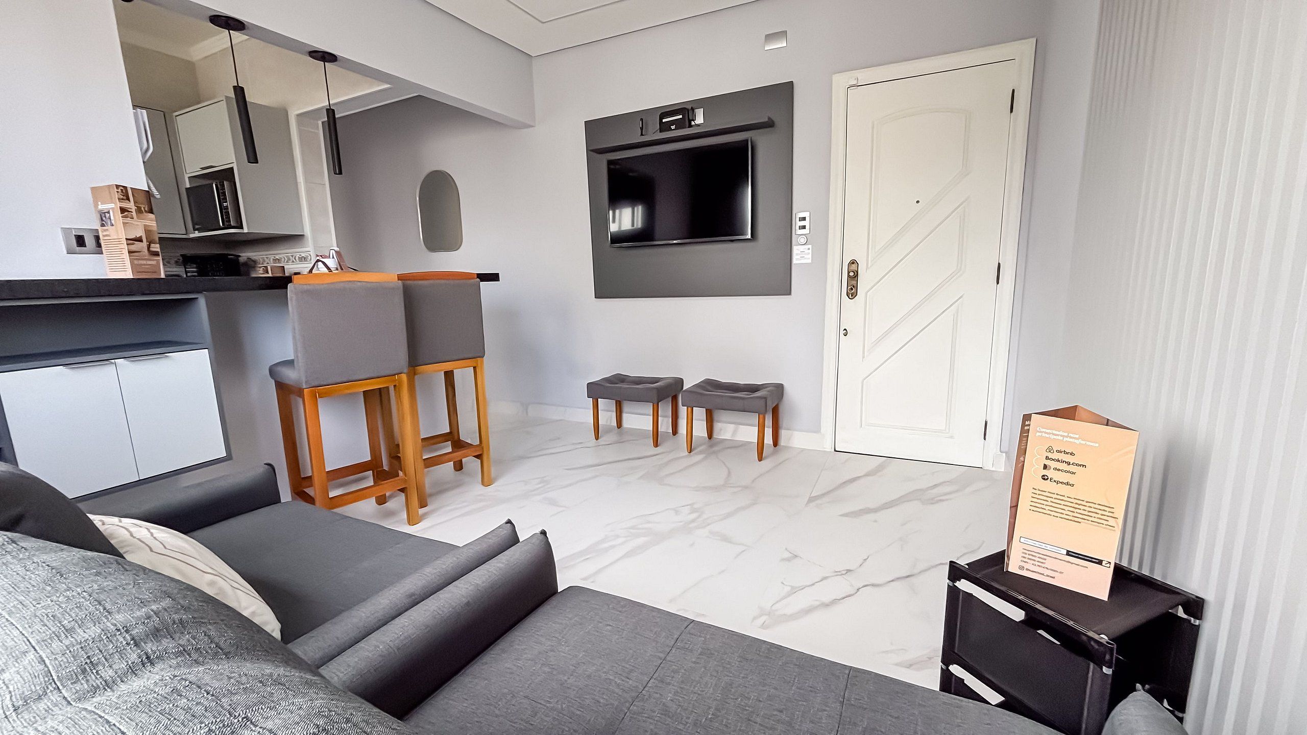 A modern living room with a sofa, a dining area with a table and chairs, a TV mounted on the wall, and a door.