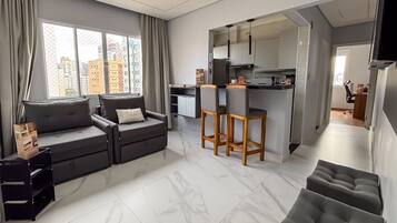 Una sala de estar moderna con un sofá, un comedor con taburetes, una cocina con nevera y armarios, y vistas de edificios a través de amplias ventanas.