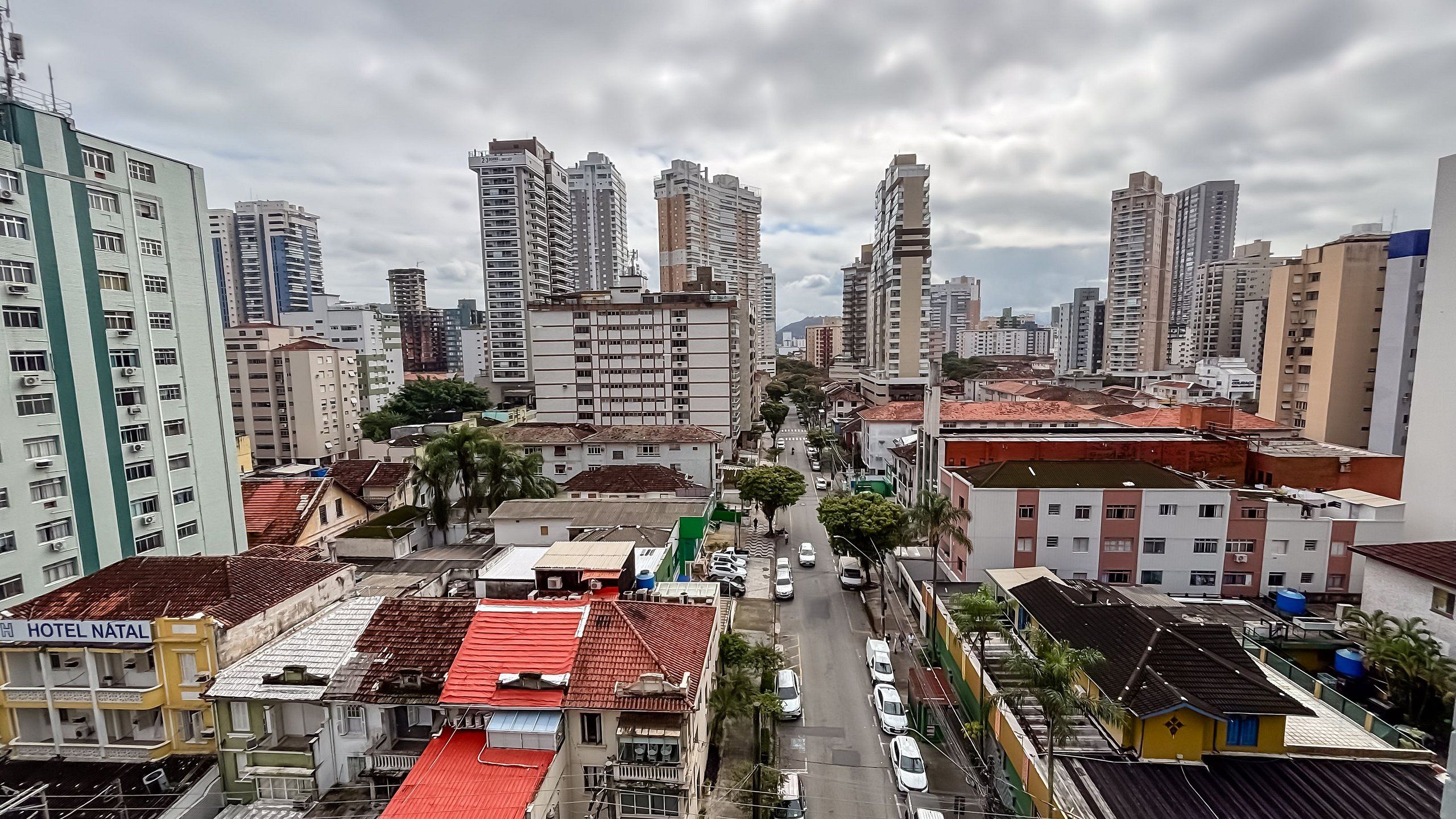 Un paysage urbain avec de hauts immeubles, une rue avec des voitures et un hôtel appelé « HÔTEL NATAL », dont le toit est rouge.