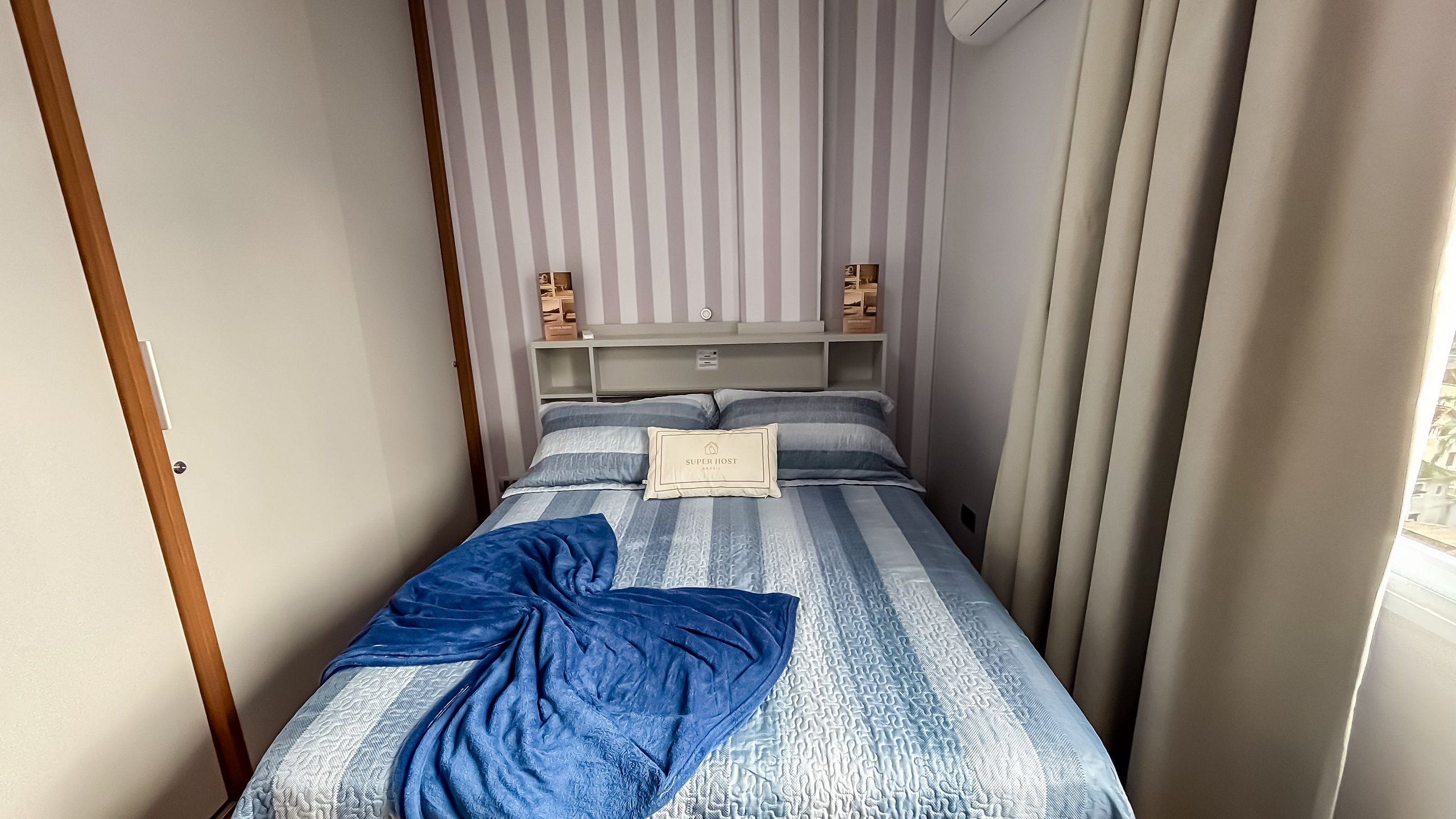 A bed with a striped bedspread, a pillow, and a blue blanket. There is a headboard with a shelf and a wall-mounted air conditioner.