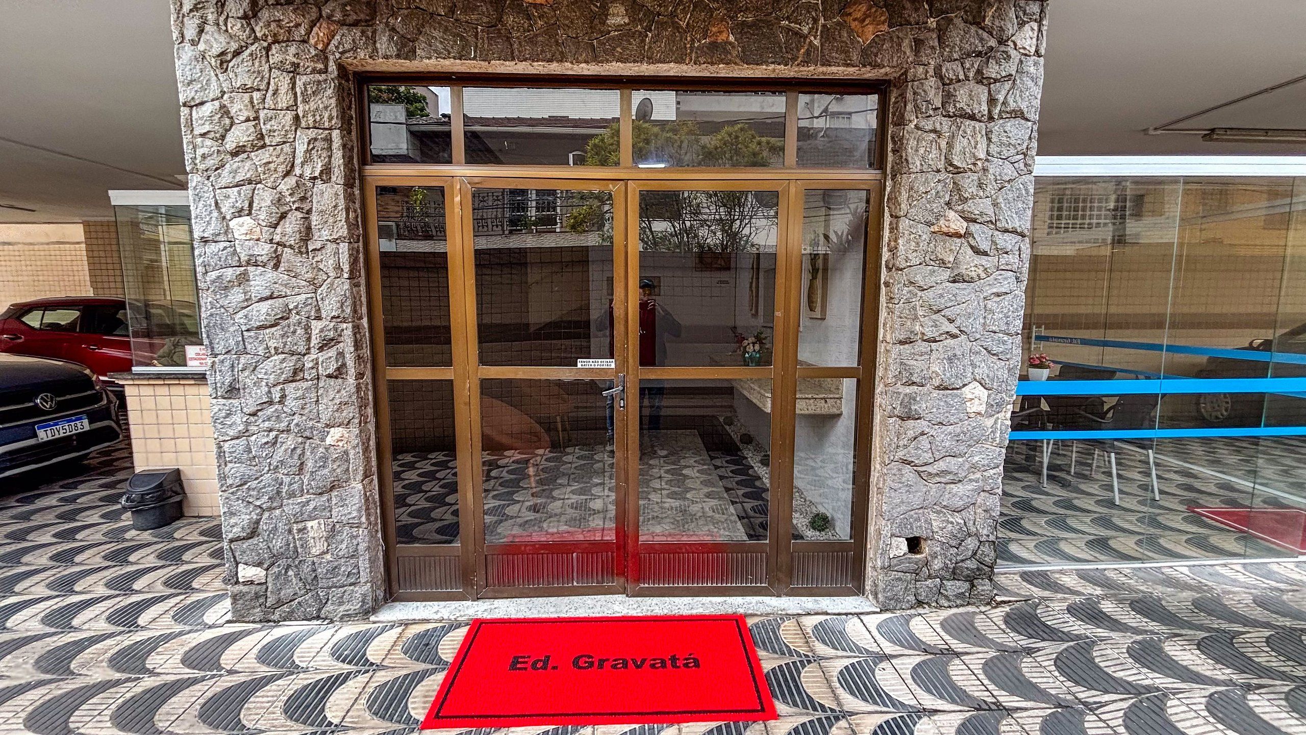 A building entrance with a stone facade, a glass door with a metal gate, and a red mat with 'Ed. Gravatá' on it.