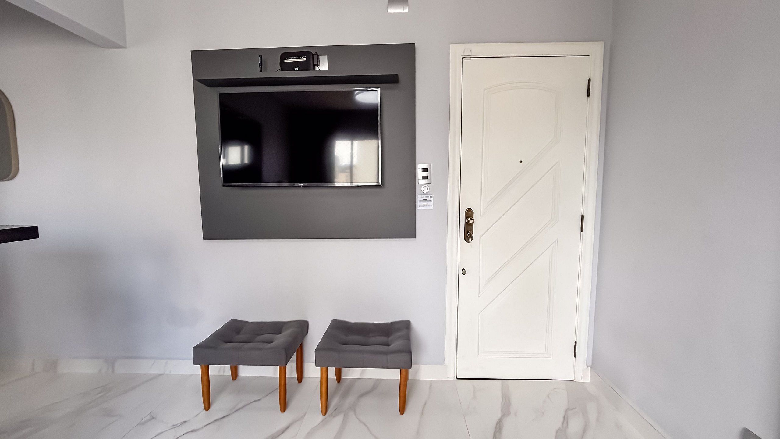 Una habitación moderna con un televisor de pantalla plana en la pared, una puerta blanca y dos taburetes grises tapizados.