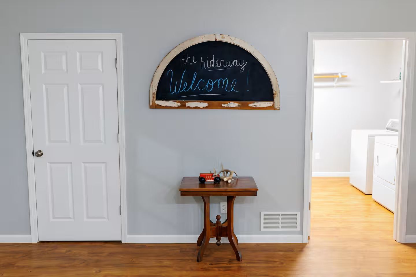A hallway with a white door, a small table with toys, and a welcome sign on the wall.