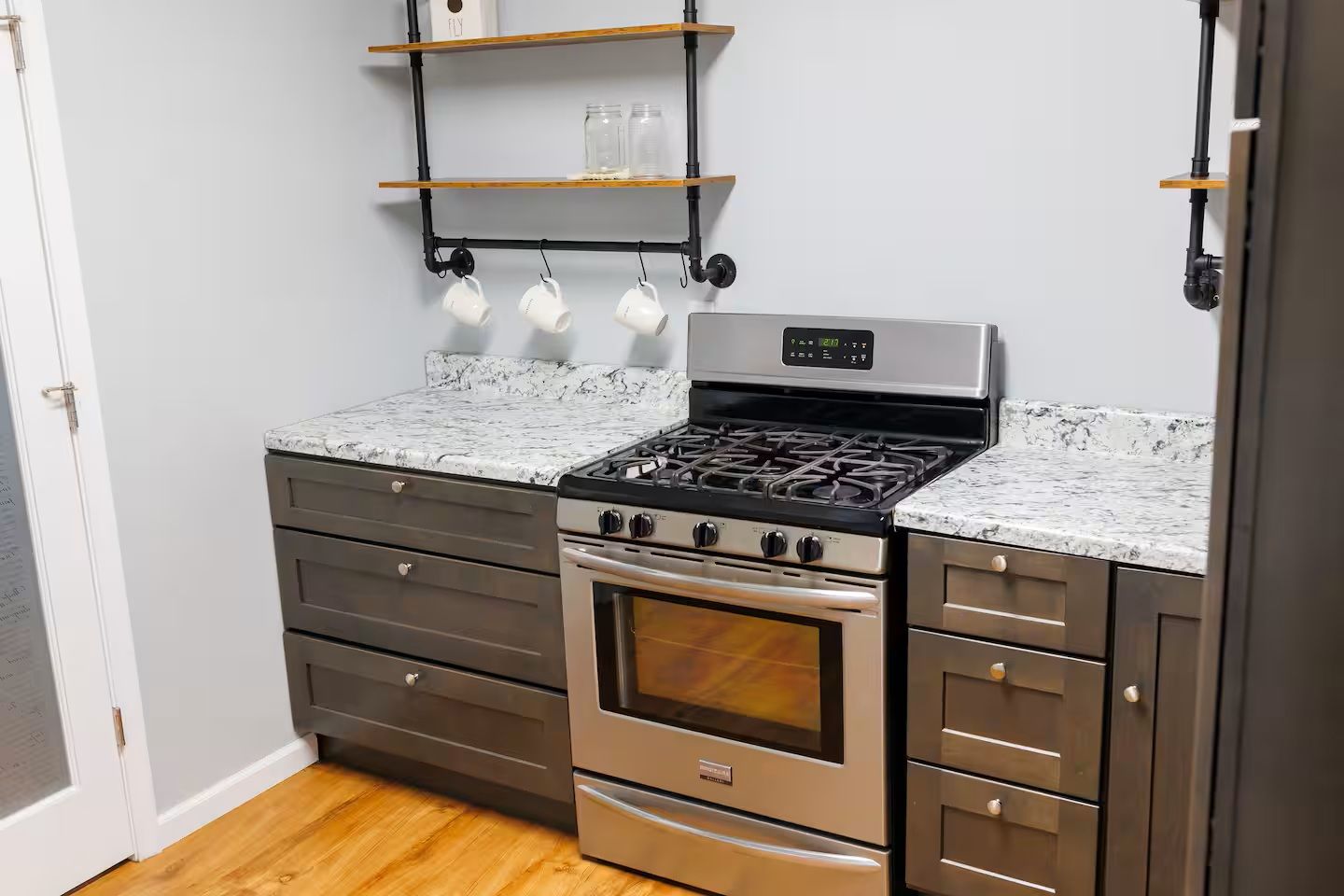 A kitchen with a gas stove, oven, and stainless steel cabinets. There are wooden shelves above the stove and a set of white mugs hanging on hooks.