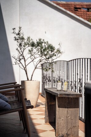 Un banc en bois, un pot de fleurs et une clôture en métal sur une terrasse dont le sol est recouvert de carreaux.