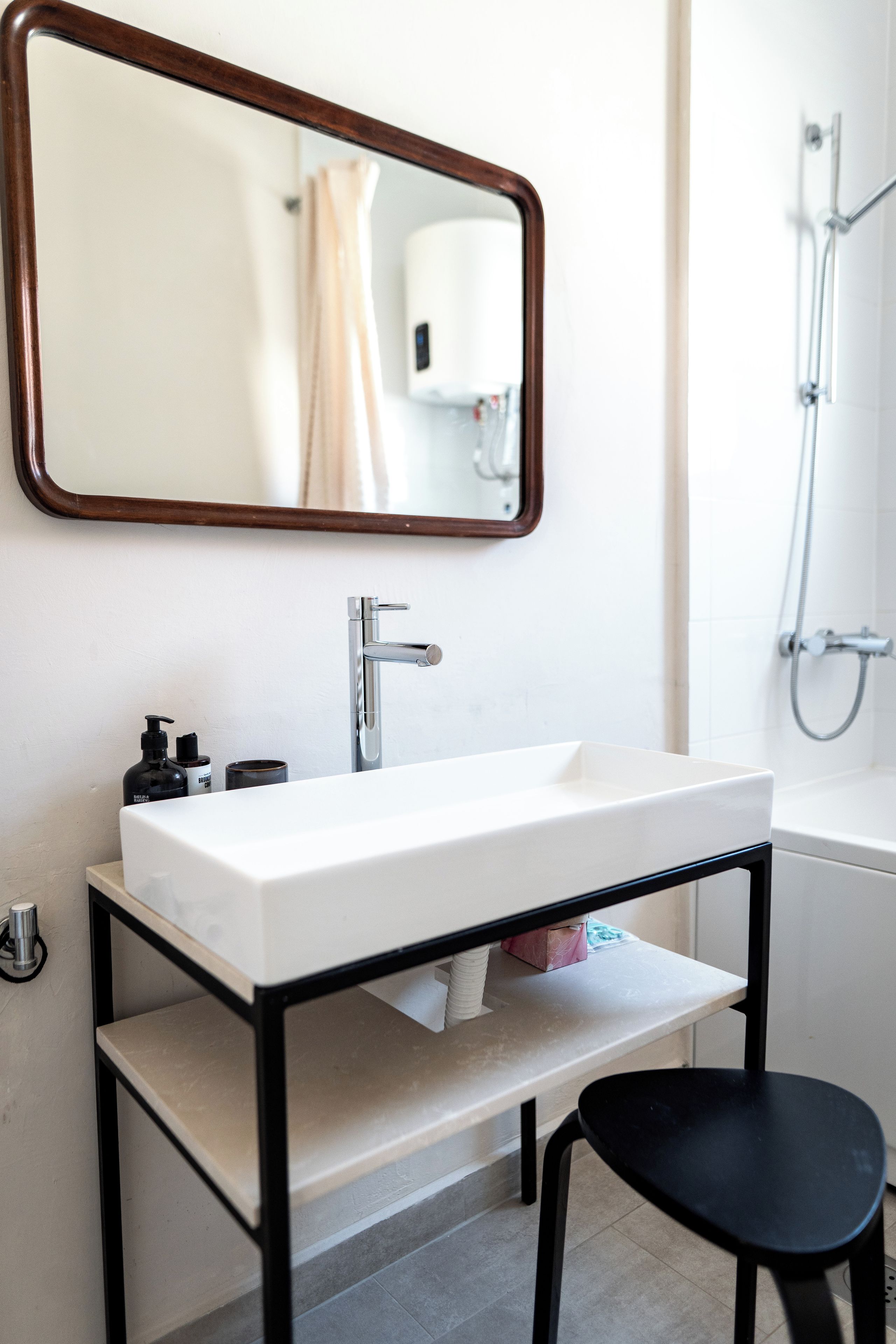 Un bagno con un lavandino bianco, uno specchio con cornice in legno, un soffione a mano e una sedia nera.