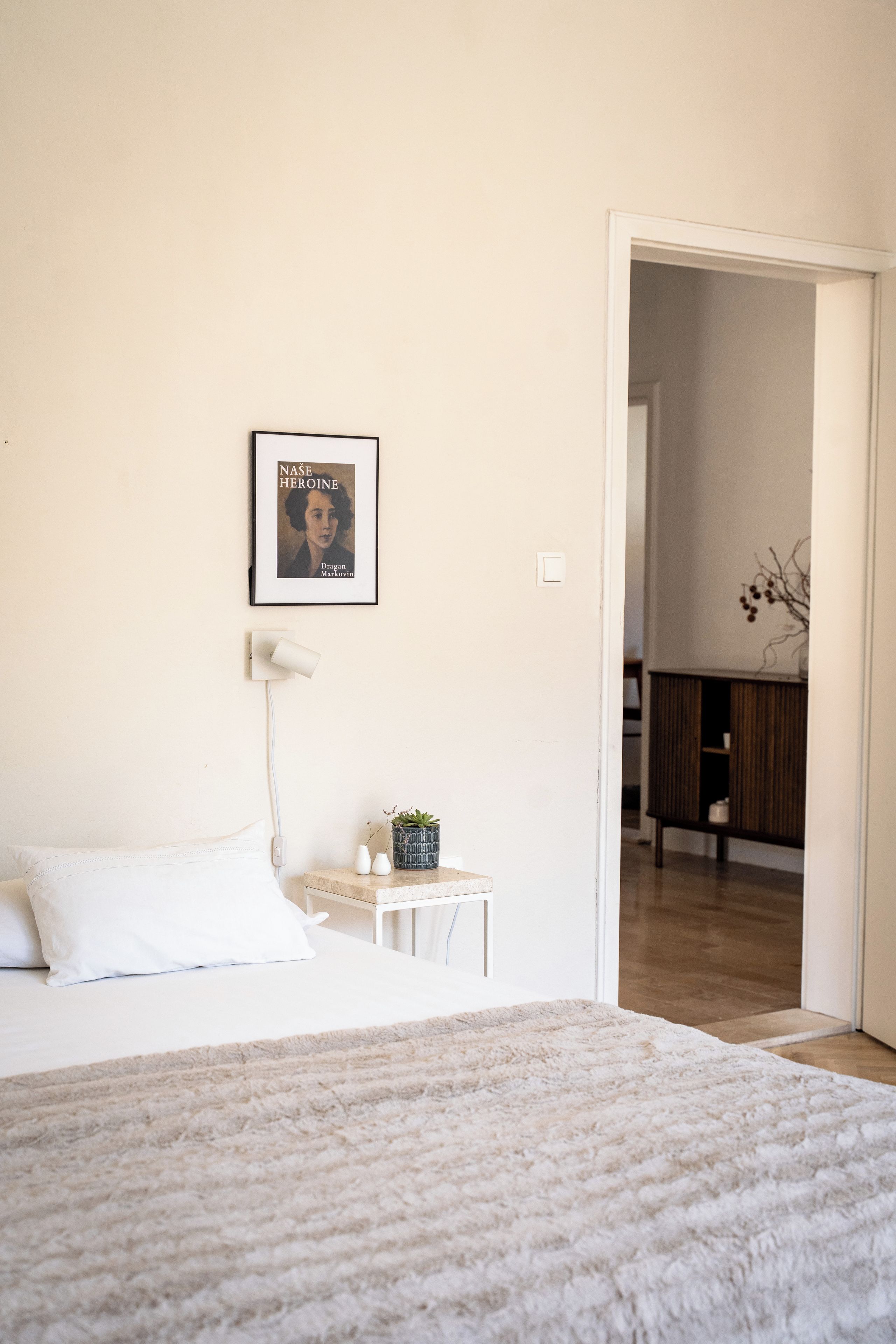 Un dormitorio con cama, una mesita de noche con lámpara, un cuadro enmarcado en la pared y un umbral que da a otra habitación.