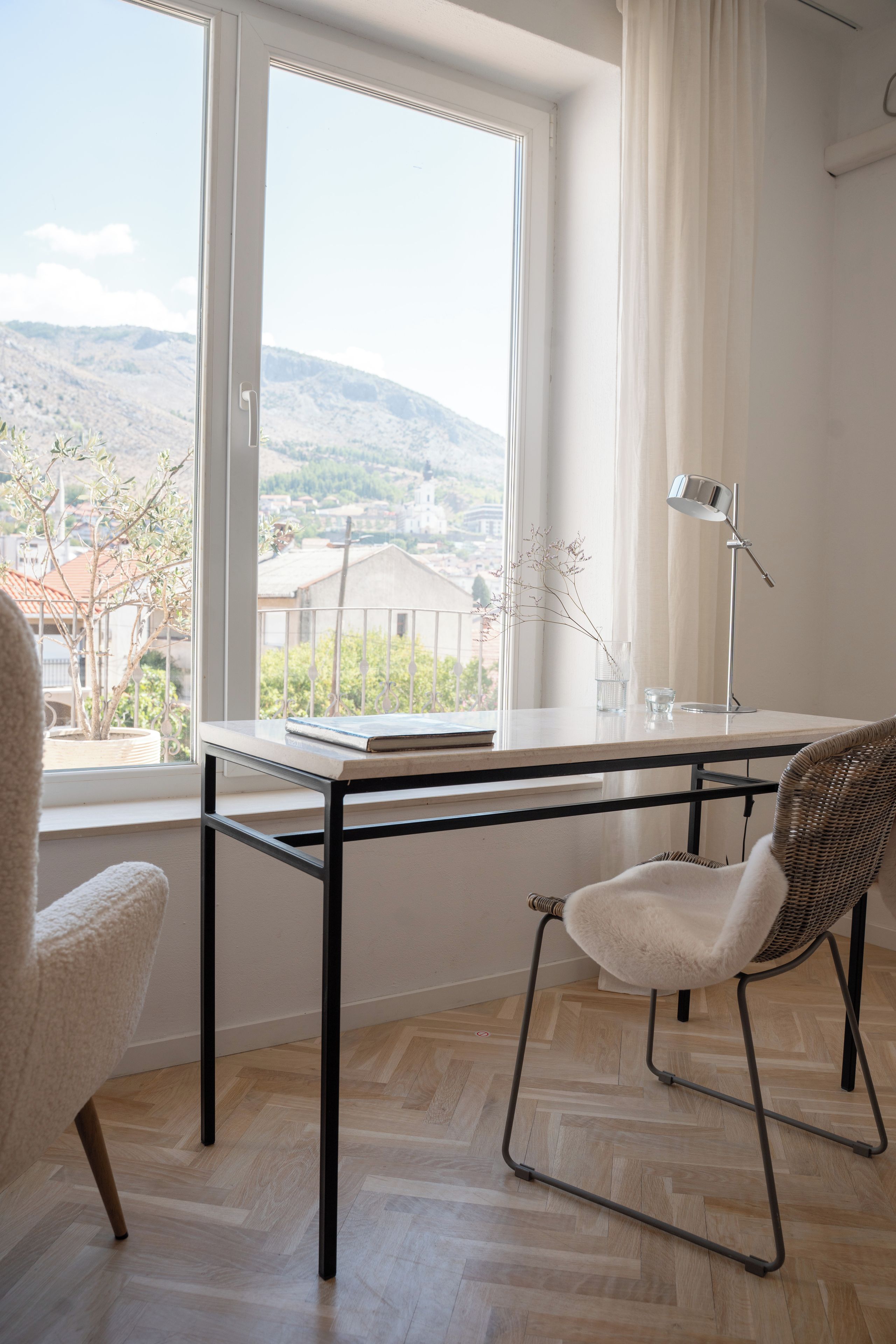 Una habitación moderna con vistas a un paisaje montañoso a través de un ventanal grande, un escritorio con lámpara y una silla.