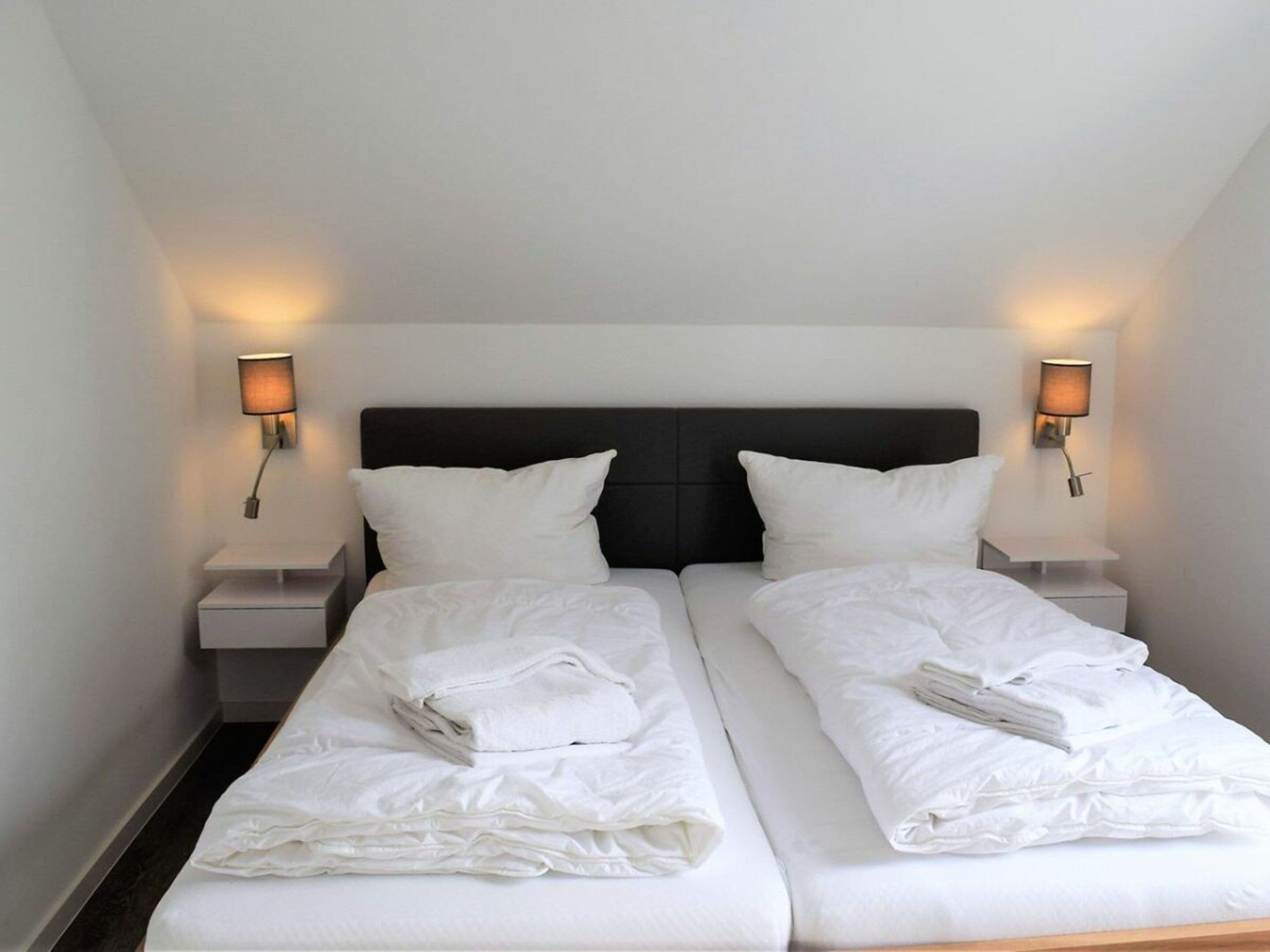 A double bed with white linens and pillows, two bedside tables, and wall-mounted lamps.