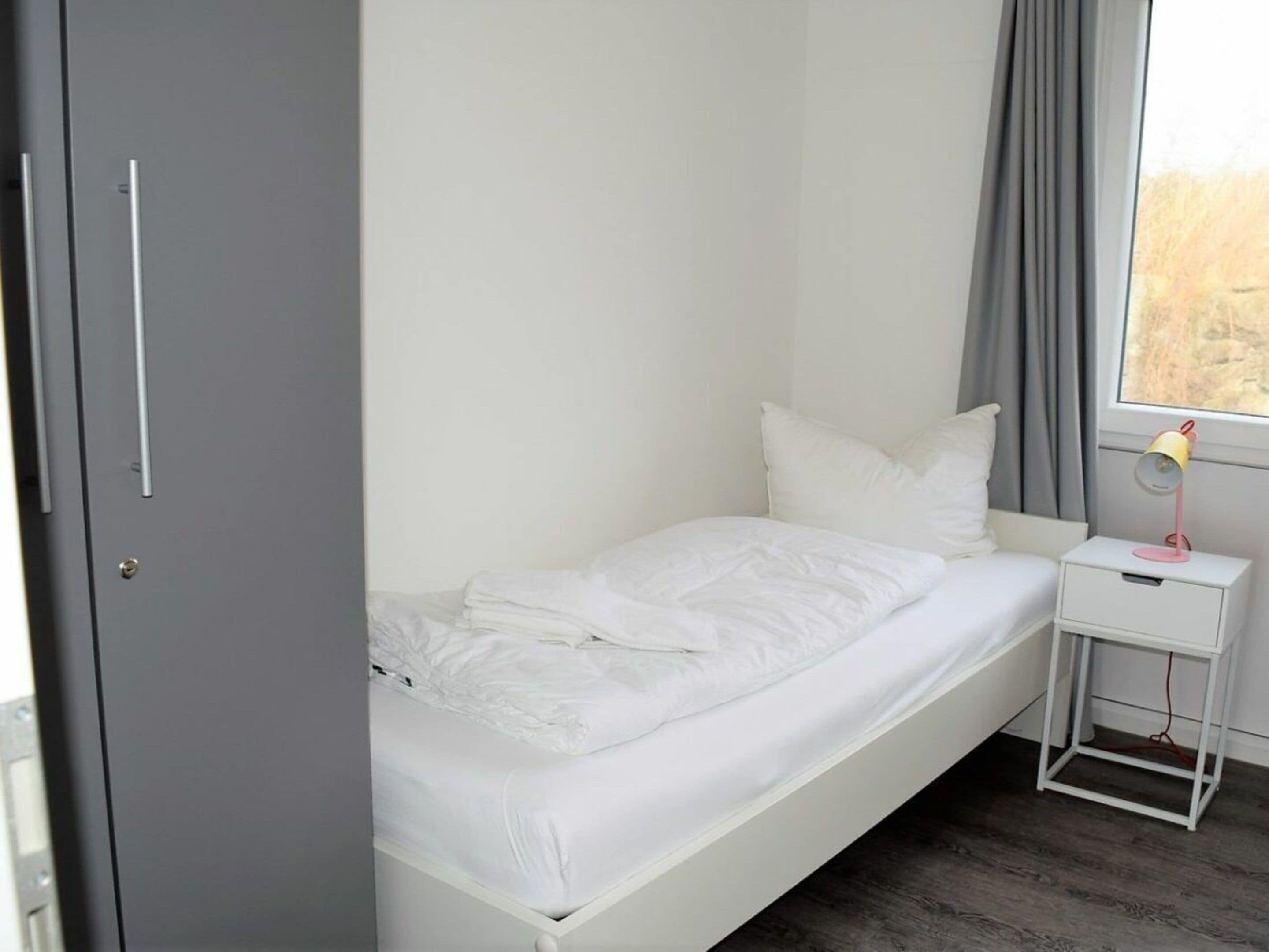 Ein Zimmer mit einem Einzelbett, einem kleinen Nachttisch mit Lampe und einem Fenster mit Vorhängen.