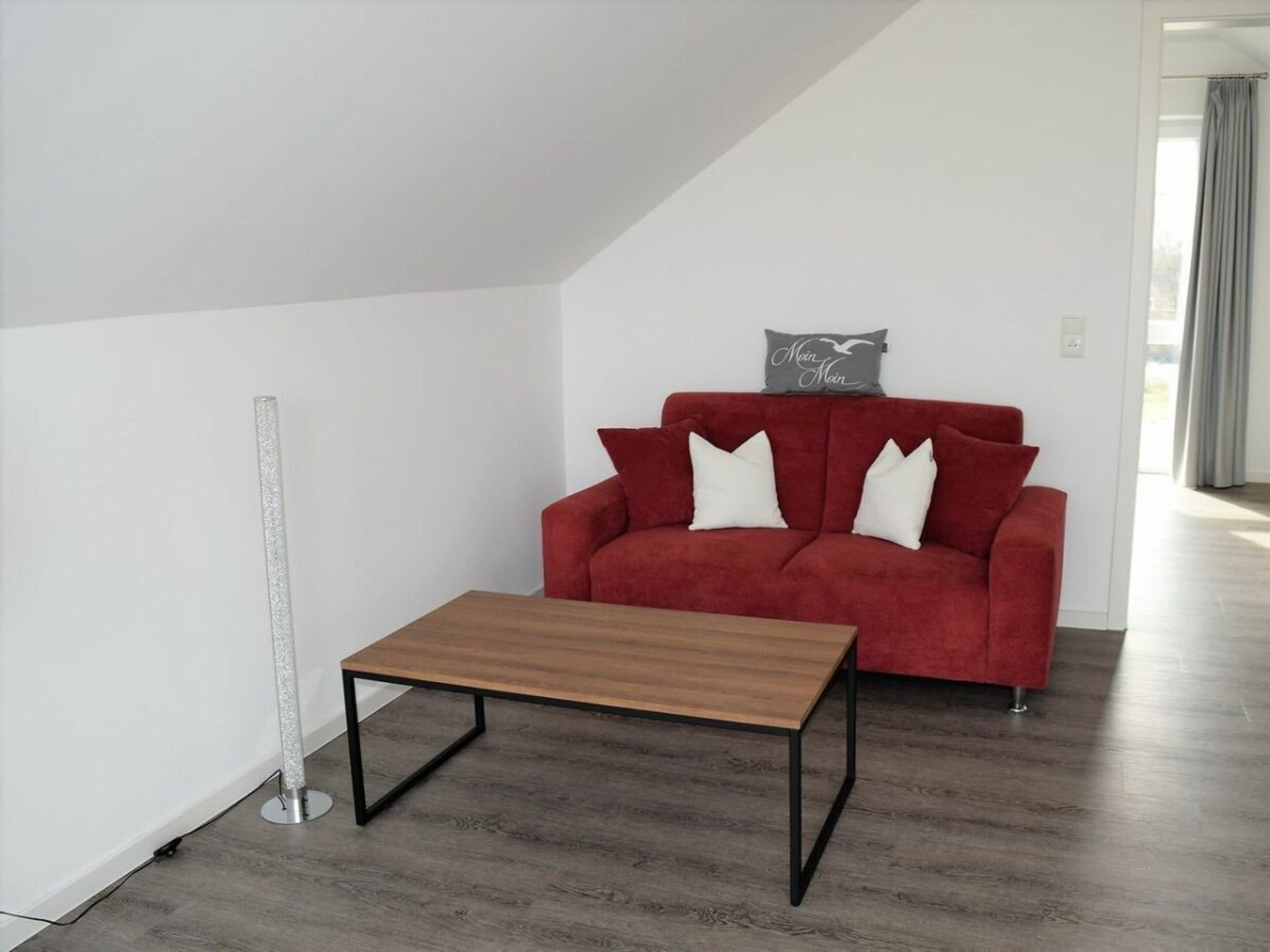 A red sofa with white cushions, a small wooden table, and a floor lamp in a room with white walls and wooden flooring.