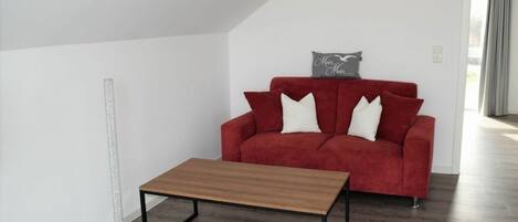 Un sofá rojo con cojines blancos, una mesita de madera y una lámpara de pie en una habitación con paredes blancas y suelo de madera.