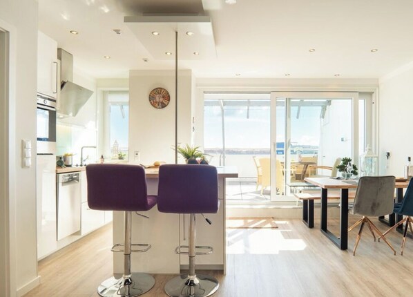 Een moderne keuken met twee barkrukken, een eethoek met een tafel en stoelen, en een grote schuifpui naar een buitenbalkon.