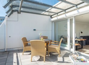 Ein moderner Außenbereich mit Korbstühlen und einem Tisch, eine Glastür, die zu einem Innenraum führt, und ein klarer Himmel.
