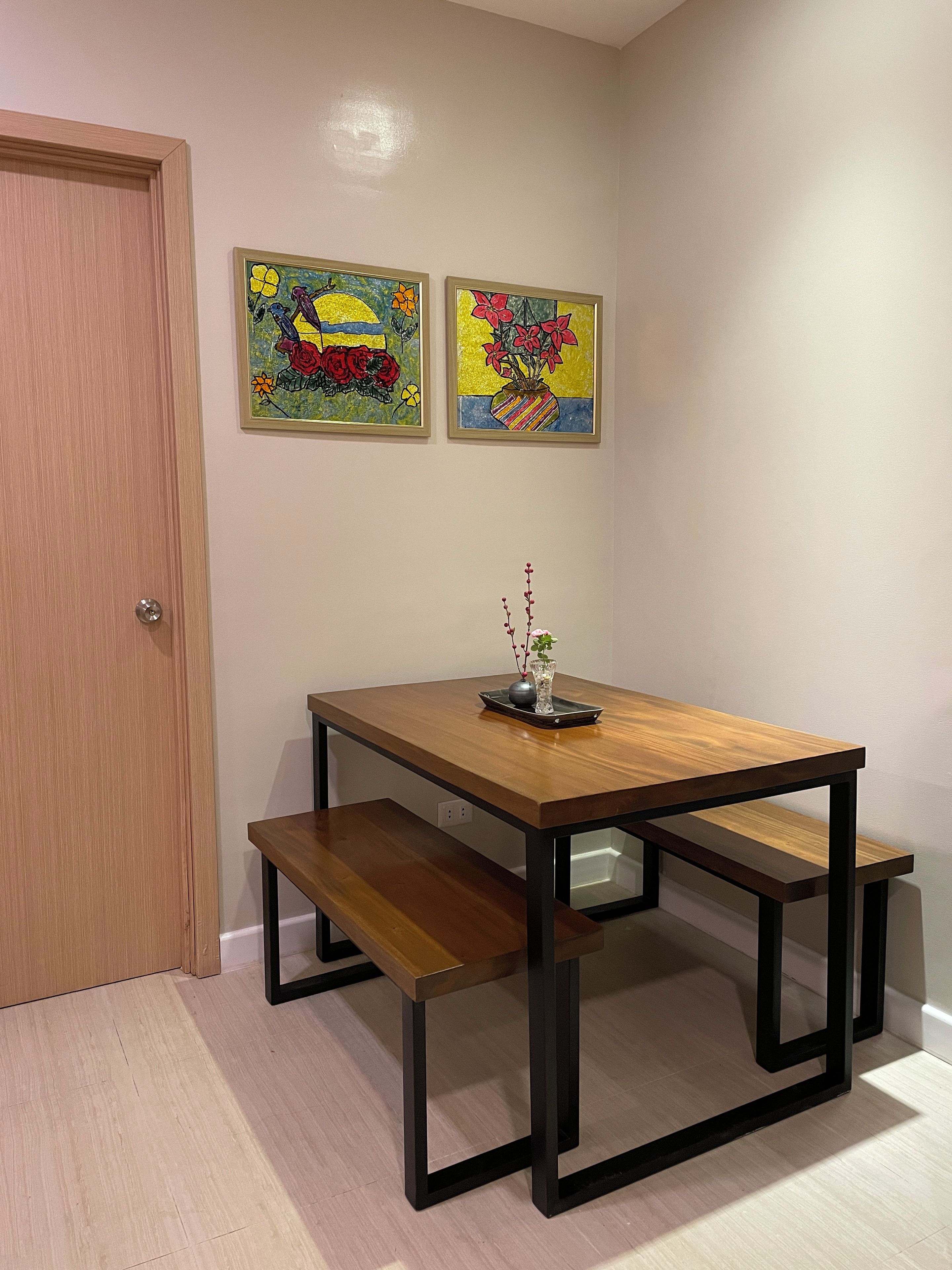 A dining area with a wooden table and benches, a vase with flowers, and two colorful paintings on the wall.
