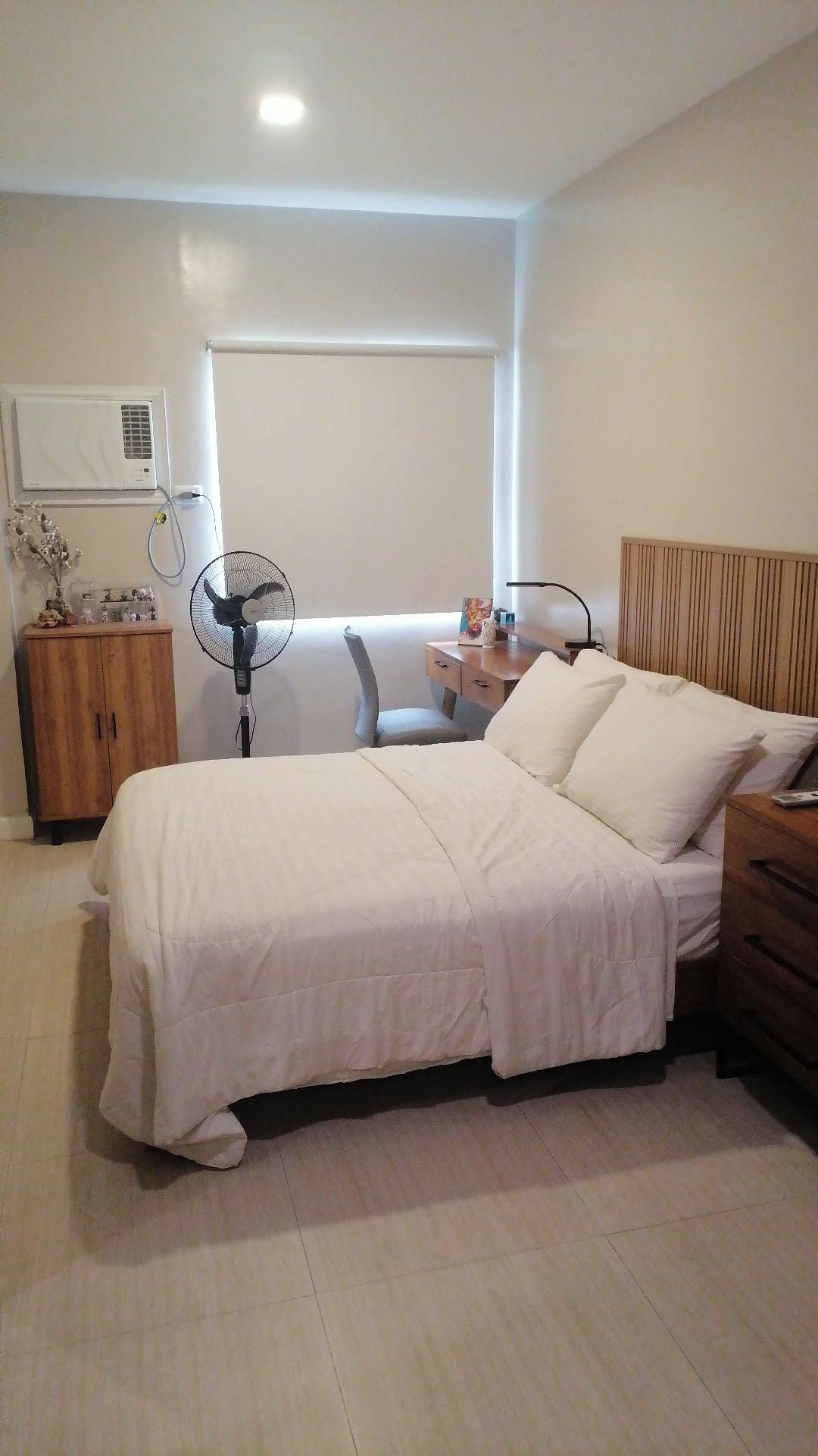 A bedroom with a bed, bedside tables, a wooden cabinet, a standing fan, and an air conditioning unit.