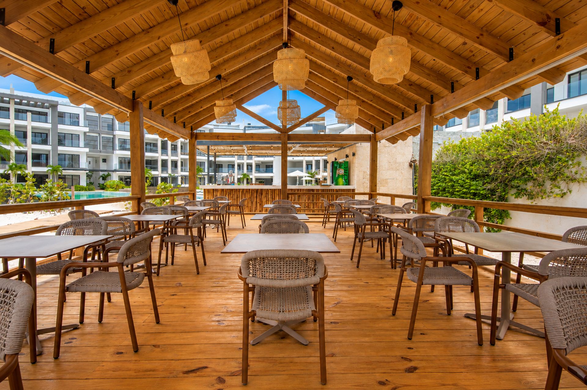 An outdoor dining area with wooden tables and chairs, a bar, and a view of buildings and greenery.