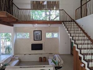 A spacious living area with a staircase, a flat-screen TV, and a large window overlooking greenery. - Casa Natik - A Mayan House in the Jungle. Ideal for families and ecotourism (Chemuyil (jungle))