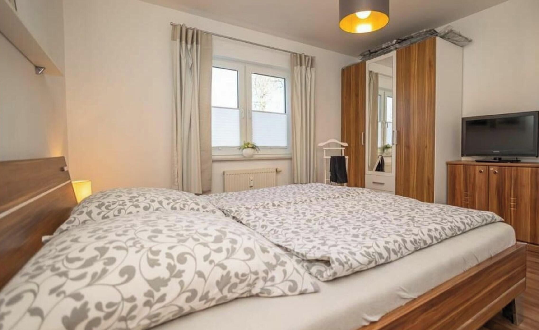 A bedroom with a double bed, a television on a wooden cabinet, a window with curtains, and a radiator.