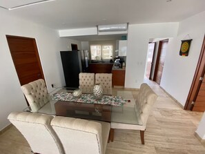 Un coin repas avec une table en verre et des chaises rembourrées, une cuisine à l’arrière-plan et un couloir menant à une porte.