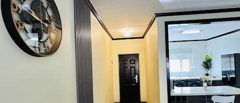 A hallway with a clock, a door, and a kitchen area with a sink and chair.