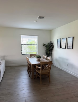 A dining area with a wooden table and chairs, a potted plant, and a window with blinds.