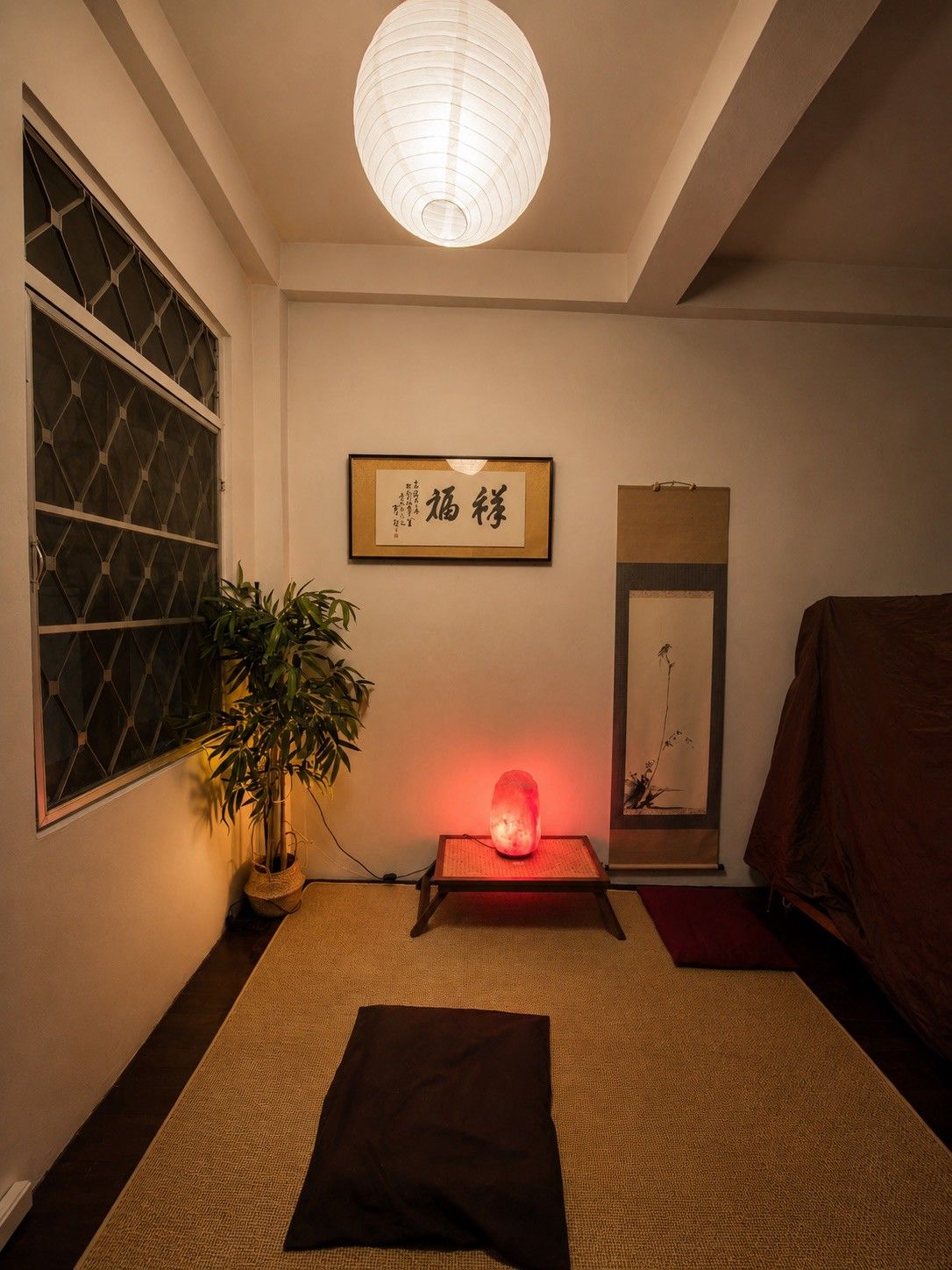A room with a traditional Japanese aesthetic, featuring a tatami mat floor, a shoji screen, a hanging paper lantern, a heated stone, and a framed calligraphy piece.