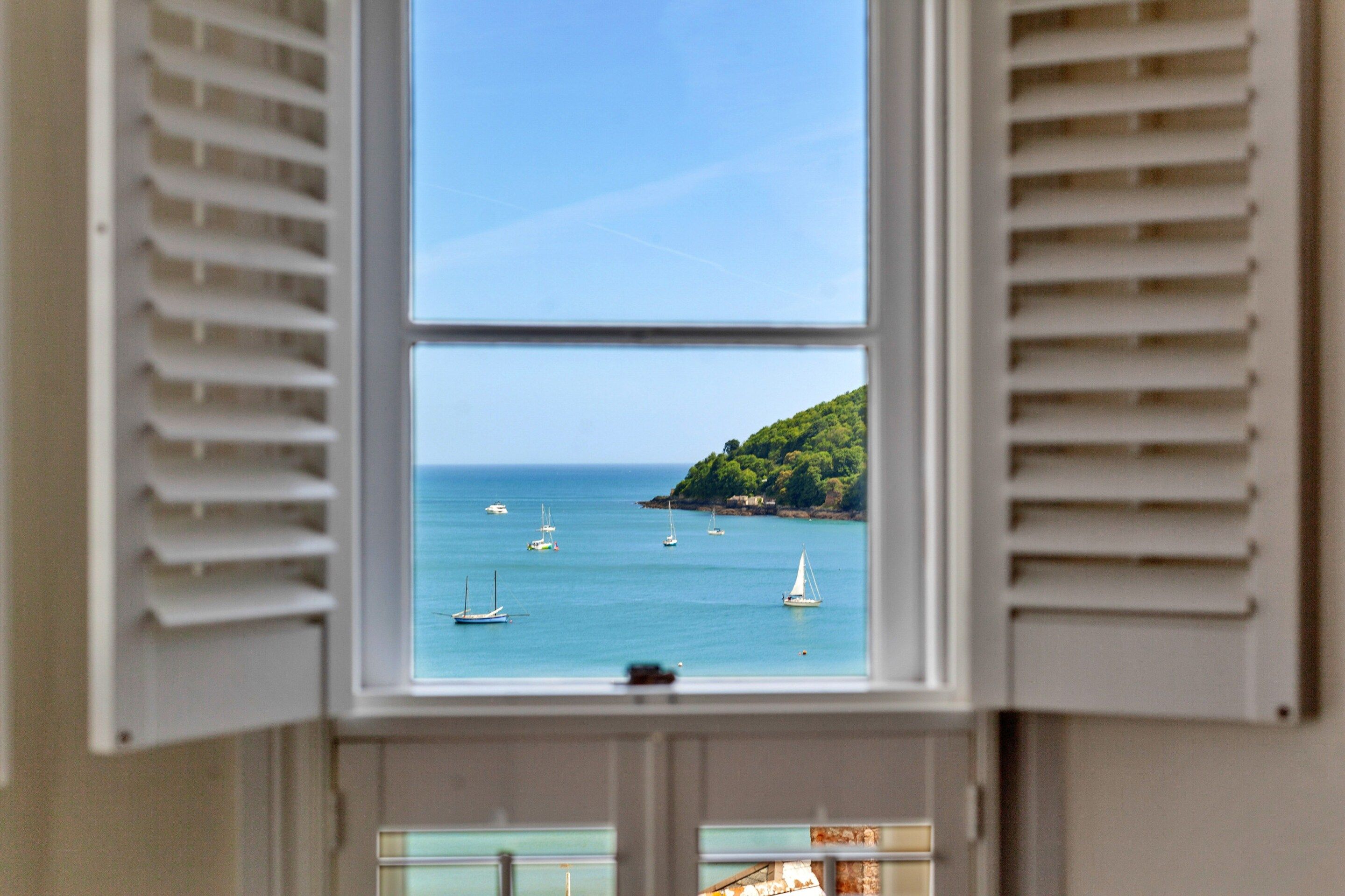 Ein Fenster mit geöffneten Rollläden, das Blick auf das Meer mit Booten und einen grünen Hügel bietet.