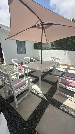 Een terras buiten met een tafel, stoelen, een grote parasol en een wit gebouw op de achtergrond.