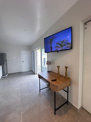 A hallway with a flat-screen TV mounted on the wall, a wooden side table with a lamp, and a door leading to an outdoor area. - Serene 5-bedroom house with AC in charming Willemstad (Willemstad)