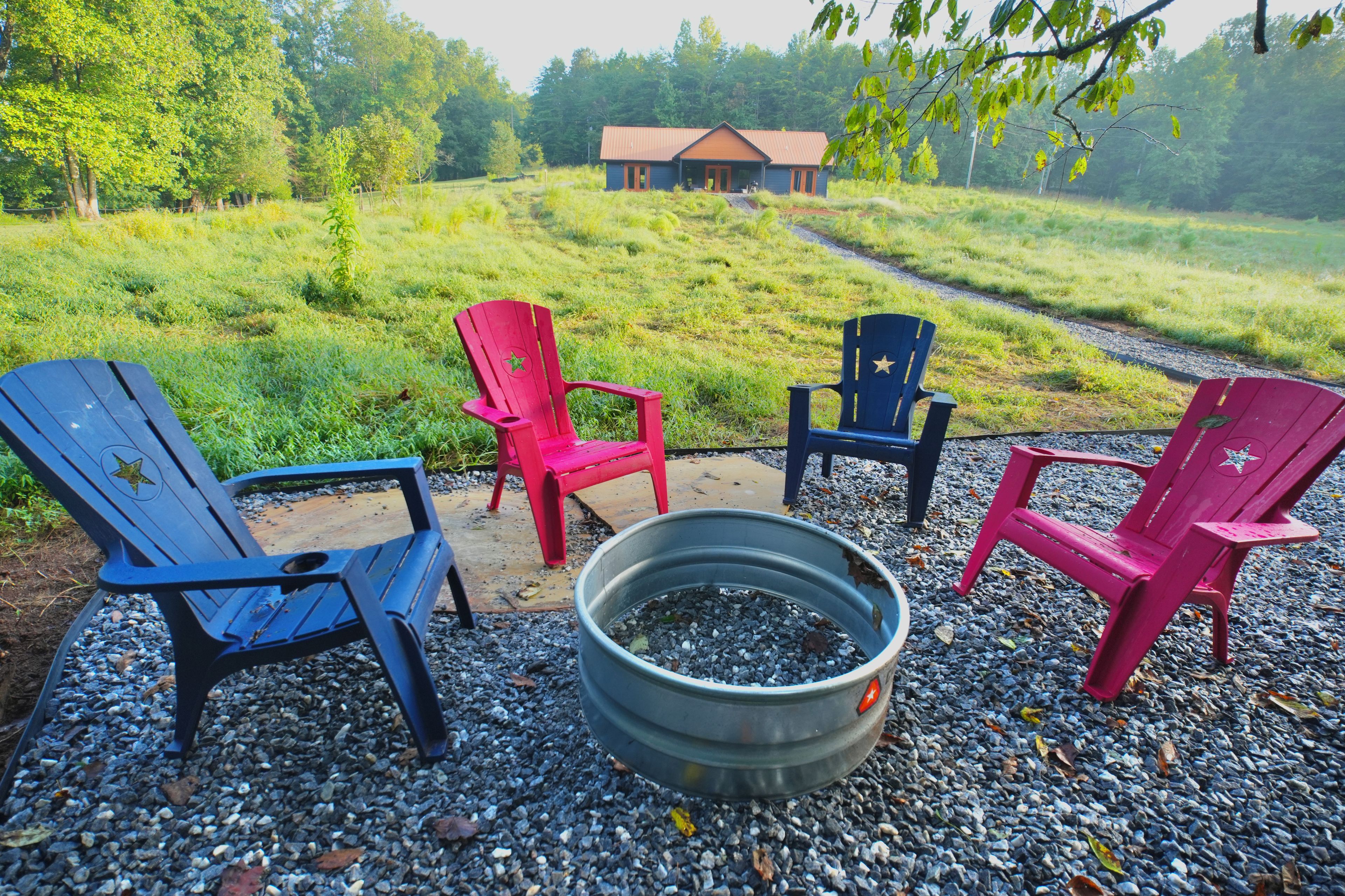 Four Adirondack chairs arranged around a metal fire pit on a gravel surface, with a wooden deck and a house in the background.