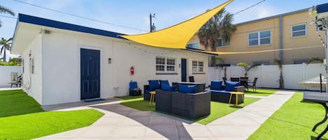 Eine moderne Außenpatio mit einem gelben Sonnensegel, ein weißes Gebäude mit einer blauen Tür und eine Sitzgruppe mit blauen Kissen.