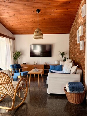 A cozy living room with a wooden ceiling, a flat-screen TV, a sofa with blue cushions, a wicker chair, and a coffee table.
