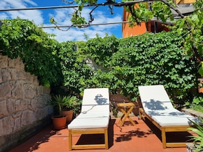 A patio with two white lounge chairs, a small wooden table, and potted plants.