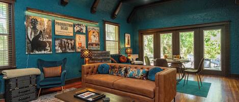 A spacious living room with a brown leather sofa, a coffee table, and a patterned rug. There are framed posters on the walls and a large window providing natural light.