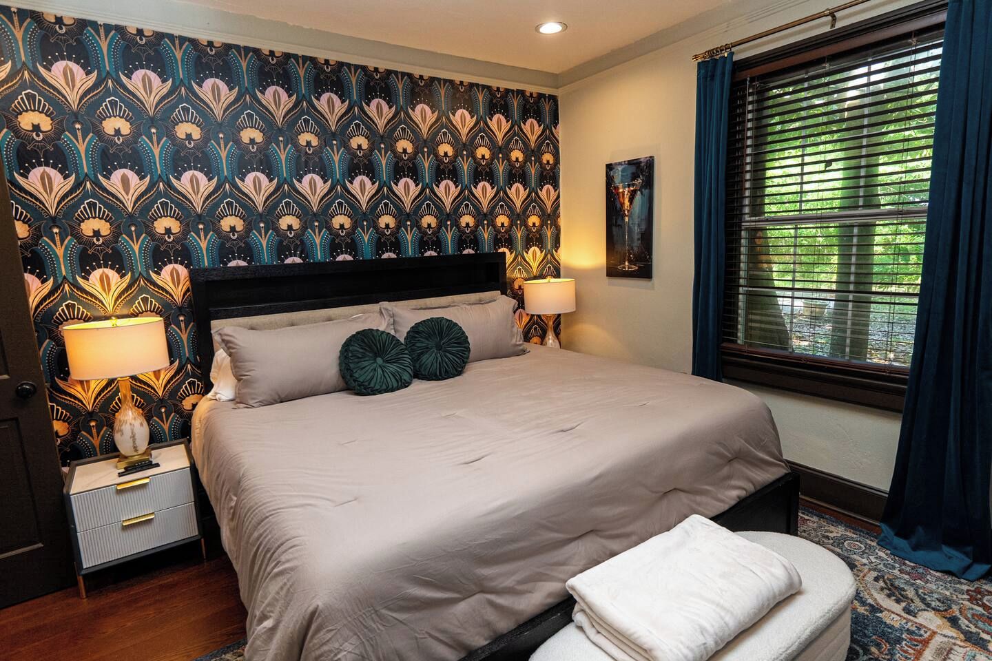 A bedroom with a patterned wallpaper, a bed with two pillows, bedside lamps, a window with blinds, and a rug.