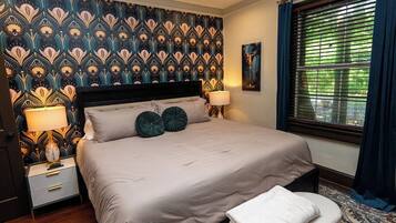 A bedroom with a patterned wallpaper, a bed with two pillows, bedside lamps, a window with blinds, and a rug.
